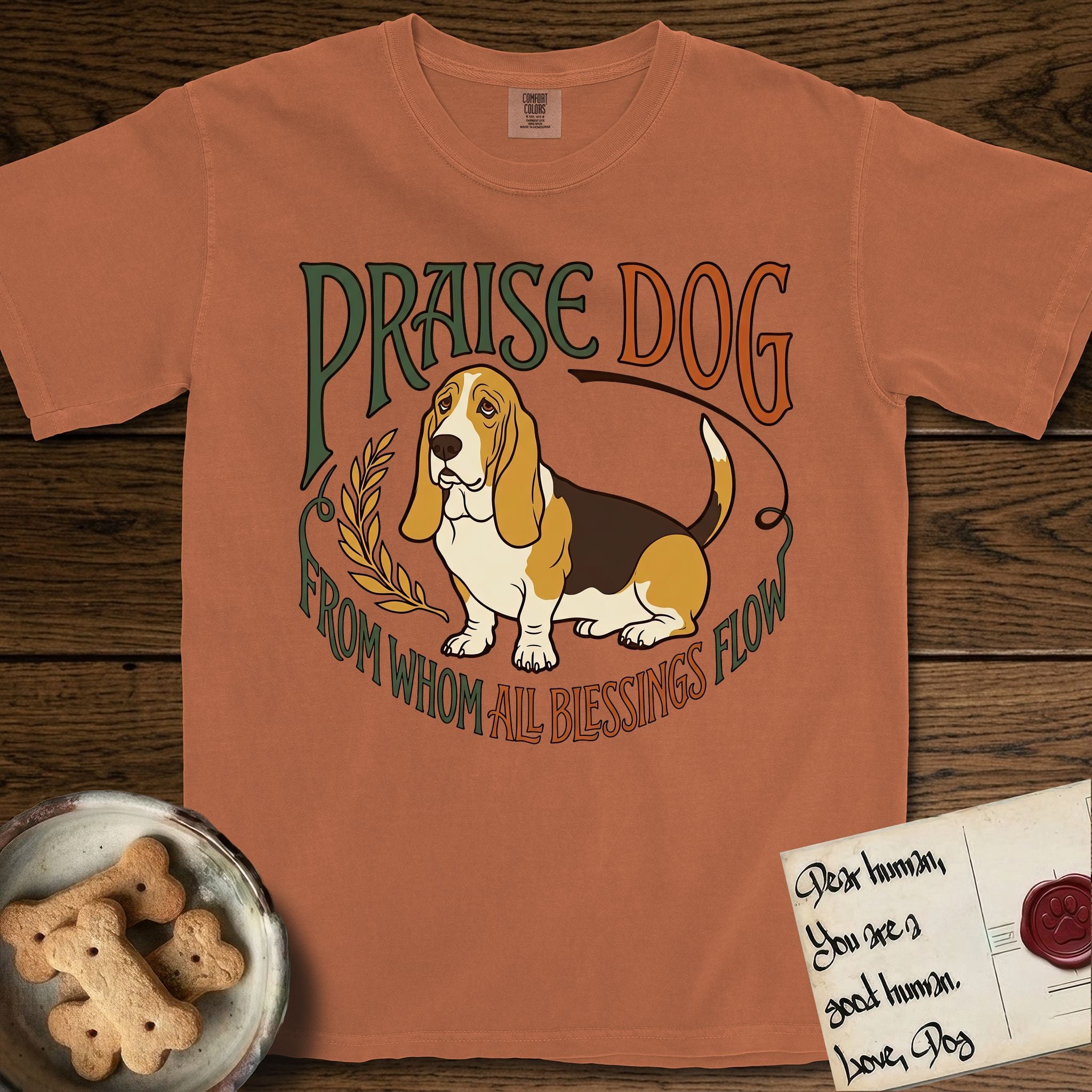 Praise Dog From Whom All Blessings Flow Basset Hound T-Shirt flat lay in Yam, featuring an art nouveau hand-lettered design with a sad-eyed Basset Hound sitting beside a golden laurel sprig and the words Praise Dog From Whom All Blessings Flow.