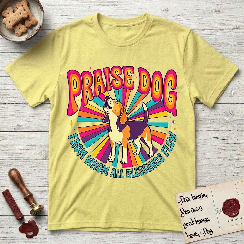 Praise Dog From Whom All Blessings Flow Beagle T-Shirt flat lay in Cornsilk, featuring a groovy psychedelic sunburst design with a Beagle howling skyward at the center and the words Praise Dog From Whom All Blessings Flow in rainbow bubble letters.