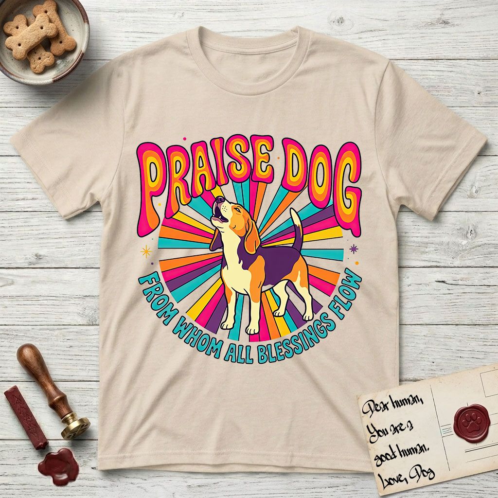 Praise Dog From Whom All Blessings Flow Beagle T-Shirt flat lay in Sand, featuring a groovy psychedelic sunburst design with a Beagle howling skyward at the center and the words Praise Dog From Whom All Blessings Flow in rainbow bubble letters.