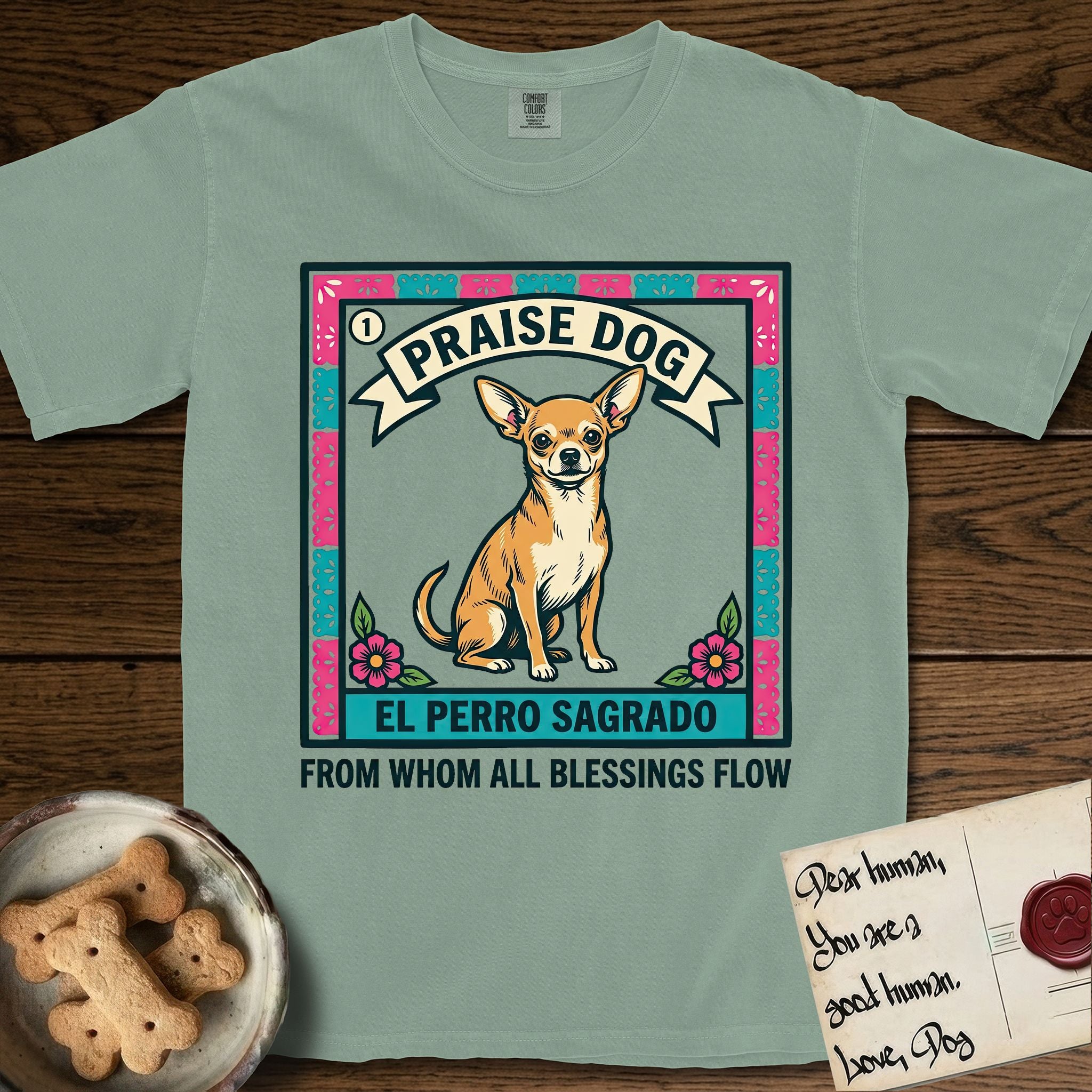Praise Dog From Whom All Blessings Flow Chihuahua T-Shirt flat lay in Bay, featuring a loteria-style folk art card with a Chihuahua seated inside a papel picado border, a banner reading El Perro Sagrado, and the words Praise Dog From Whom All Blessings Flow.