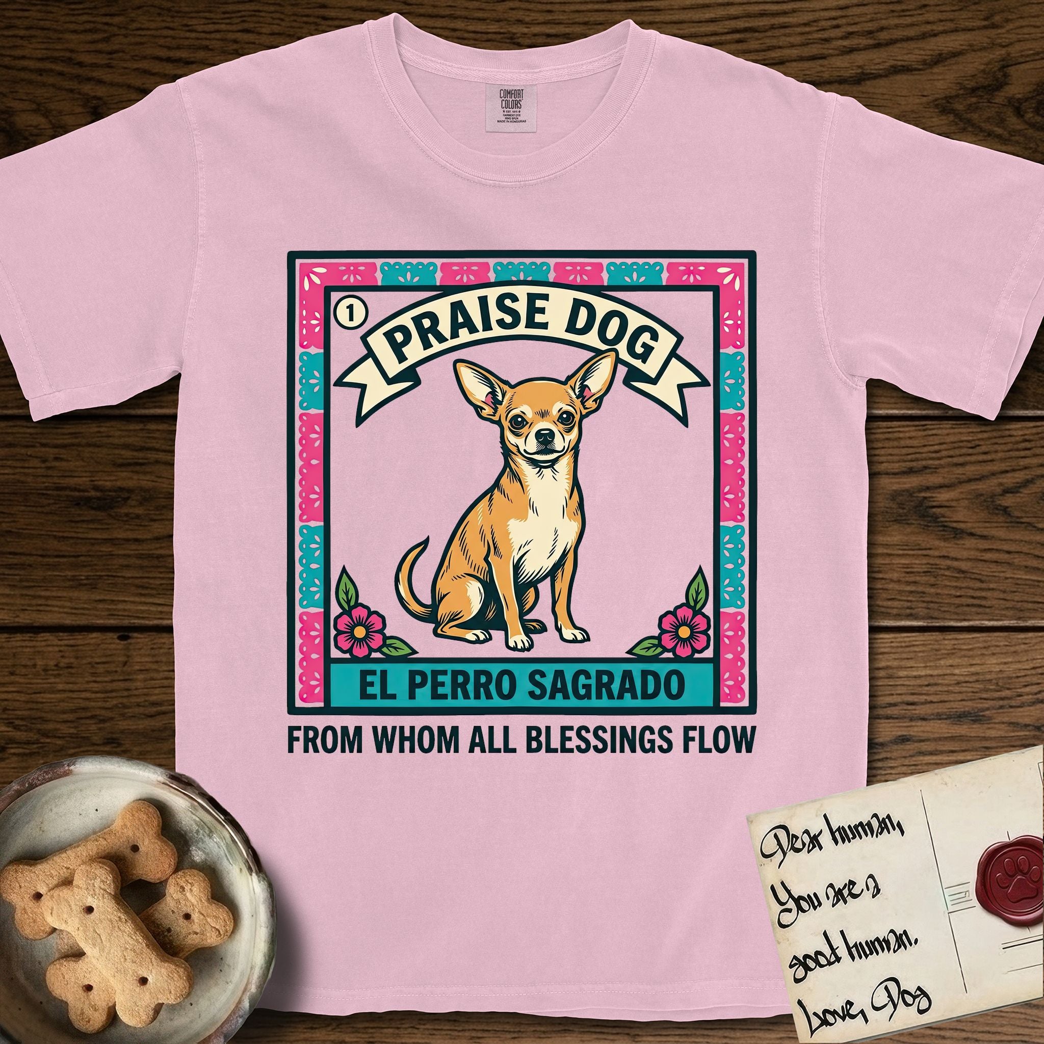 Praise Dog From Whom All Blessings Flow Chihuahua T-Shirt flat lay in Blossom, featuring a loteria-style folk art card with a Chihuahua seated inside a papel picado border, a banner reading El Perro Sagrado, and the words Praise Dog From Whom All Blessings Flow.