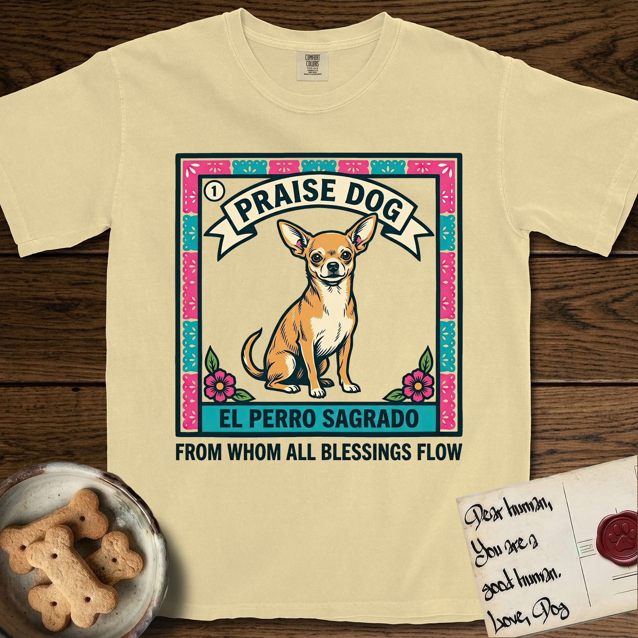 Praise Dog From Whom All Blessings Flow Chihuahua T-Shirt flat lay in Butter, featuring a loteria-style folk art card with a Chihuahua seated inside a papel picado border, a banner reading El Perro Sagrado, and the words Praise Dog From Whom All Blessings Flow.