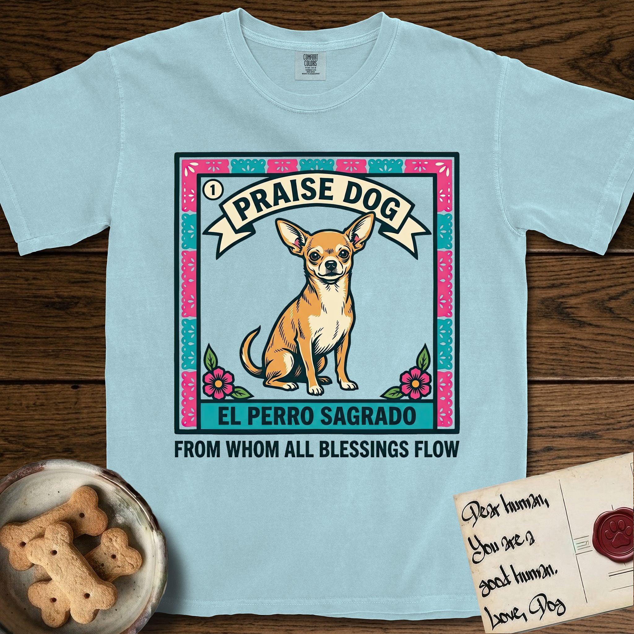 Praise Dog From Whom All Blessings Flow Chihuahua T-Shirt flat lay in Chambray, featuring a loteria-style folk art card with a Chihuahua seated inside a papel picado border, a banner reading El Perro Sagrado, and the words Praise Dog From Whom All Blessings Flow.
