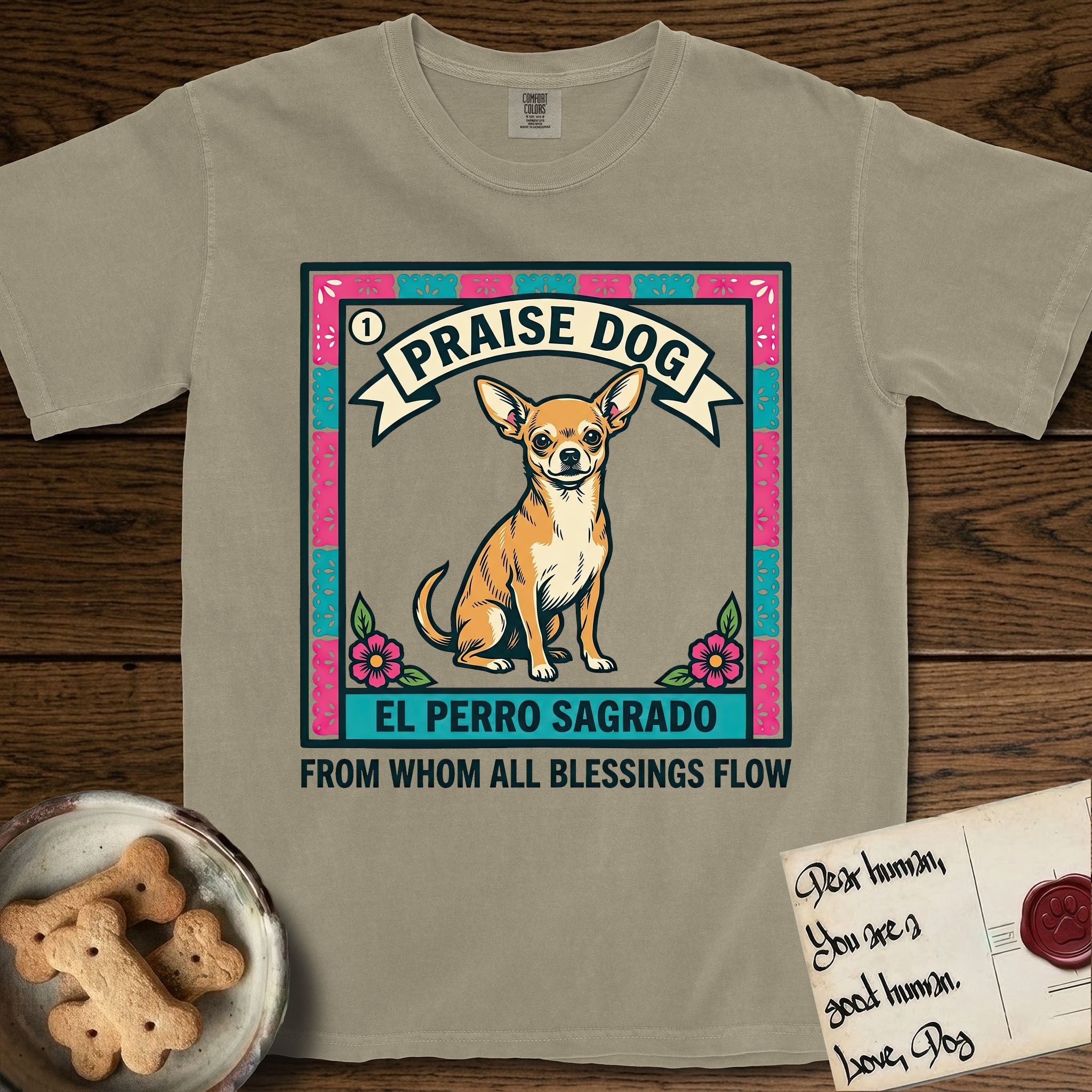 Praise Dog From Whom All Blessings Flow Chihuahua T-Shirt flat lay in Khaki, featuring a loteria-style folk art card with a Chihuahua seated inside a papel picado border, a banner reading El Perro Sagrado, and the words Praise Dog From Whom All Blessings Flow.