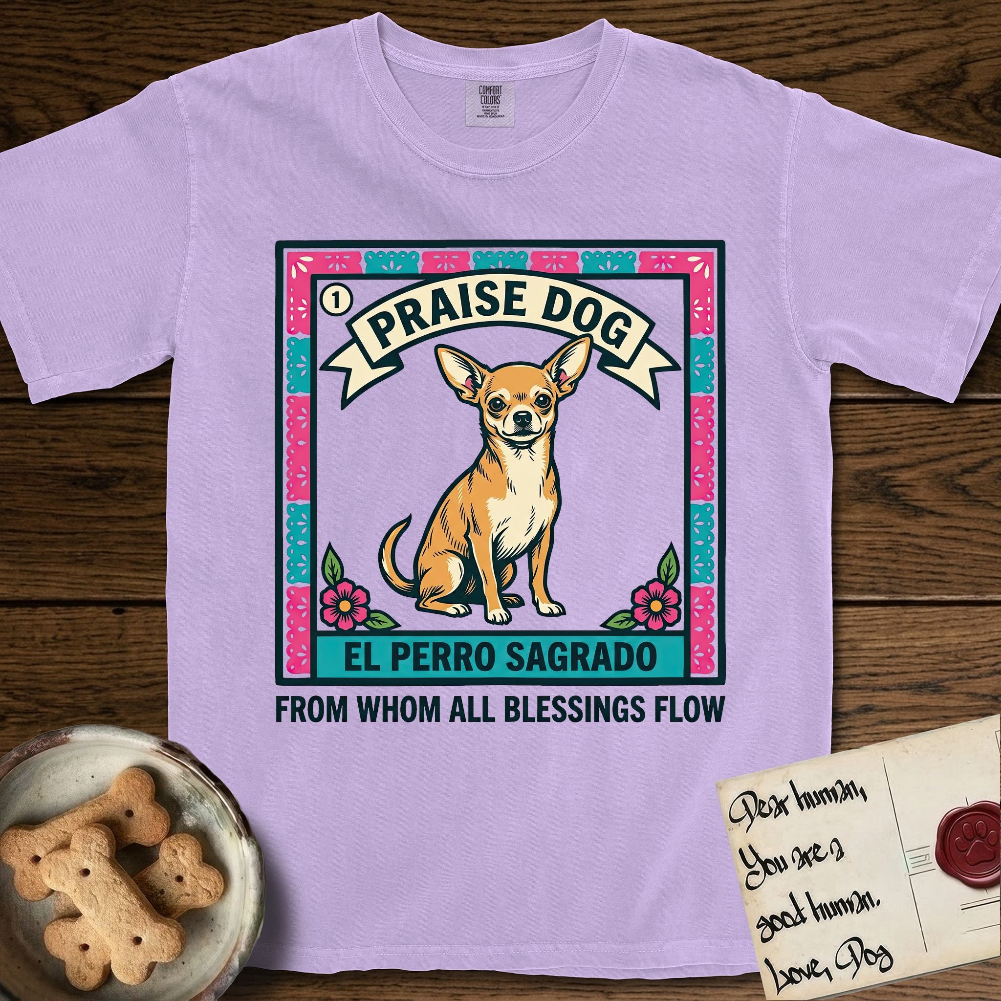 Praise Dog From Whom All Blessings Flow Chihuahua T-Shirt flat lay in Orchid, featuring a loteria-style folk art card with a Chihuahua seated inside a papel picado border, a banner reading El Perro Sagrado, and the words Praise Dog From Whom All Blessings Flow.