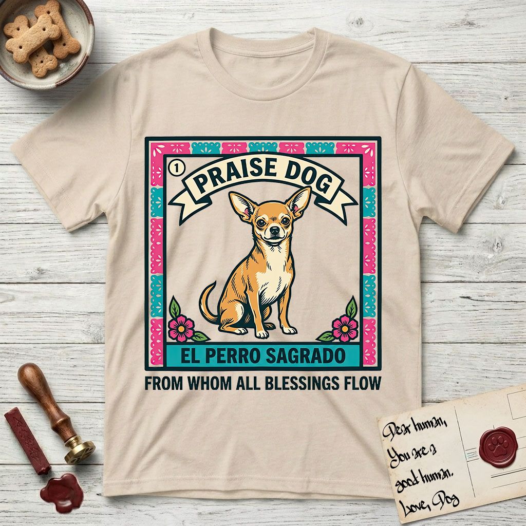 Praise Dog From Whom All Blessings Flow Chihuahua T-Shirt flat lay in Sand, featuring a loteria-style folk art card with a Chihuahua seated inside a papel picado border, a banner reading El Perro Sagrado, and the words Praise Dog From Whom All Blessings Flow.