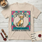 Praise Dog From Whom All Blessings Flow Chihuahua T-Shirt flat lay in Sand, featuring a loteria-style folk art card with a Chihuahua seated inside a papel picado border, a banner reading El Perro Sagrado, and the words Praise Dog From Whom All Blessings Flow.