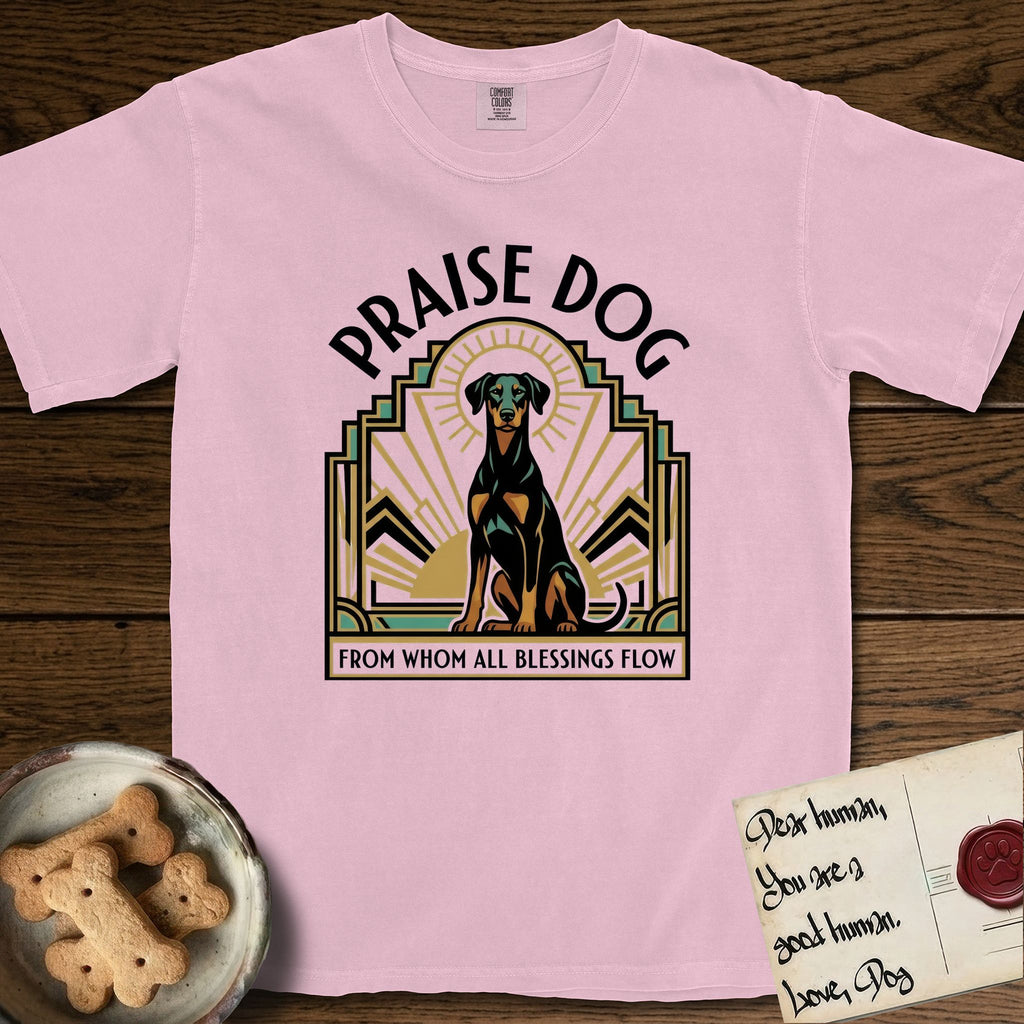 Praise Dog From Whom All Blessings Flow Doberman T-Shirt flat lay in Blossom, featuring an Art Deco emblem of a seated Doberman with natural ears and full tail, framed by geometric gold and jade rays with a sunburst halo and "Praise Dog From Whom All Blessings Flow" lettering.