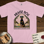 Praise Dog From Whom All Blessings Flow Doberman T-Shirt flat lay in Blossom, featuring an Art Deco emblem of a seated Doberman with natural ears and full tail, framed by geometric gold and jade rays with a sunburst halo and "Praise Dog From Whom All Blessings Flow" lettering.