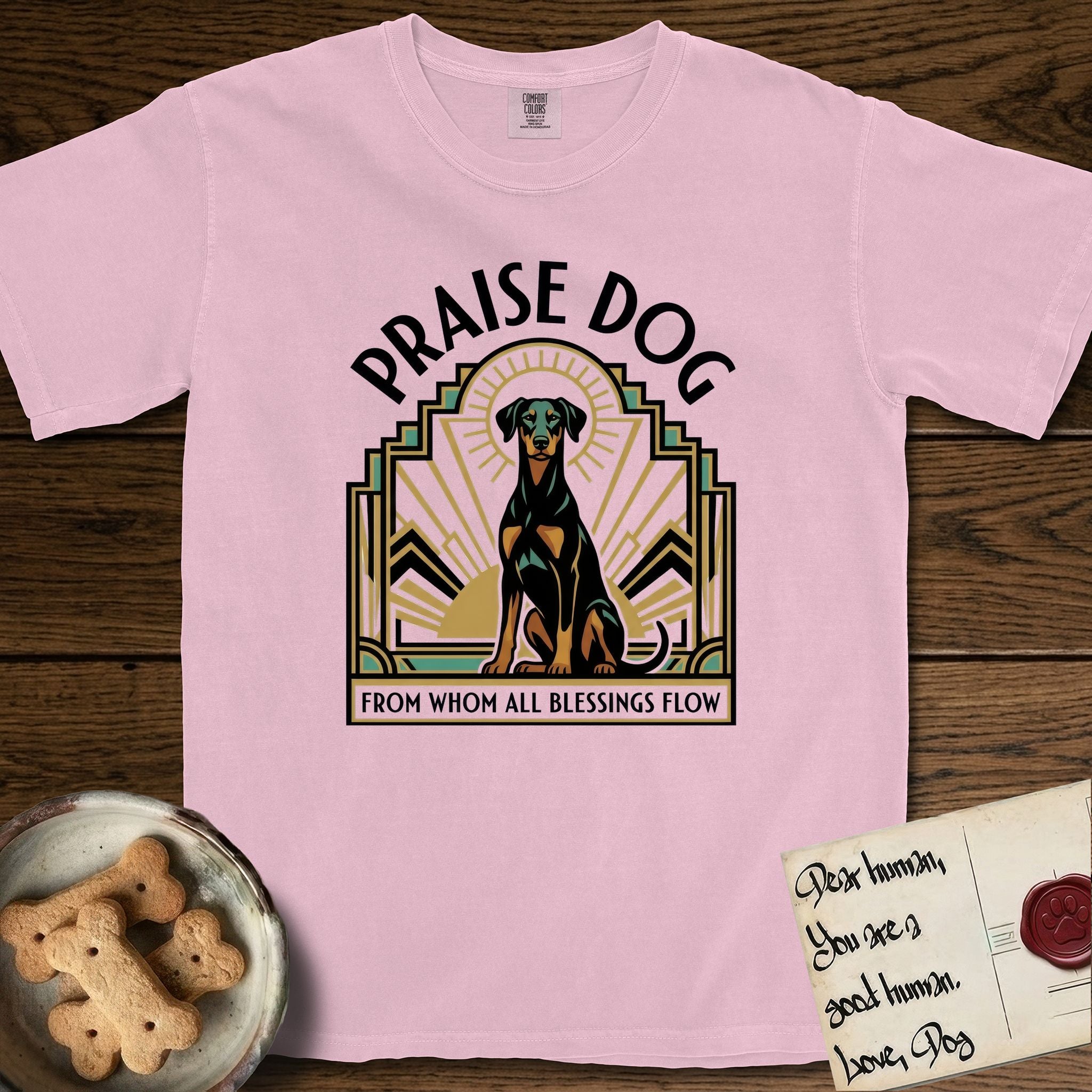 Praise Dog From Whom All Blessings Flow Doberman T-Shirt flat lay in Blossom, featuring an Art Deco emblem of a seated Doberman with natural ears and full tail, framed by geometric gold and jade rays with a sunburst halo and "Praise Dog From Whom All Blessings Flow" lettering.