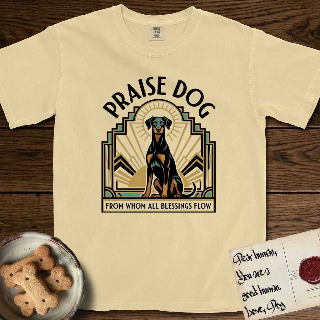 Praise Dog From Whom All Blessings Flow Doberman T-Shirt flat lay in Butter, featuring an Art Deco emblem of a seated Doberman with natural ears and full tail, framed by geometric gold and jade rays with a sunburst halo and "Praise Dog From Whom All Blessings Flow" lettering.