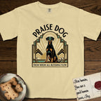 Praise Dog From Whom All Blessings Flow Doberman T-Shirt flat lay in Butter, featuring an Art Deco emblem of a seated Doberman with natural ears and full tail, framed by geometric gold and jade rays with a sunburst halo and "Praise Dog From Whom All Blessings Flow" lettering.