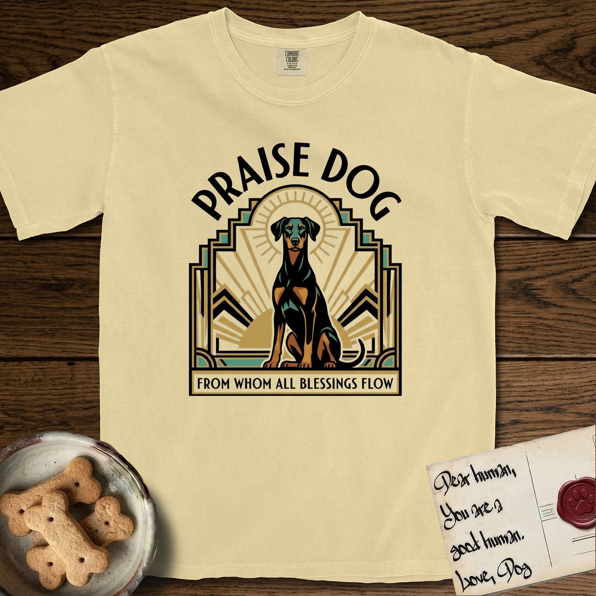 Praise Dog From Whom All Blessings Flow Doberman T-Shirt flat lay in Butter, featuring an Art Deco emblem of a seated Doberman with natural ears and full tail, framed by geometric gold and jade rays with a sunburst halo and "Praise Dog From Whom All Blessings Flow" lettering.