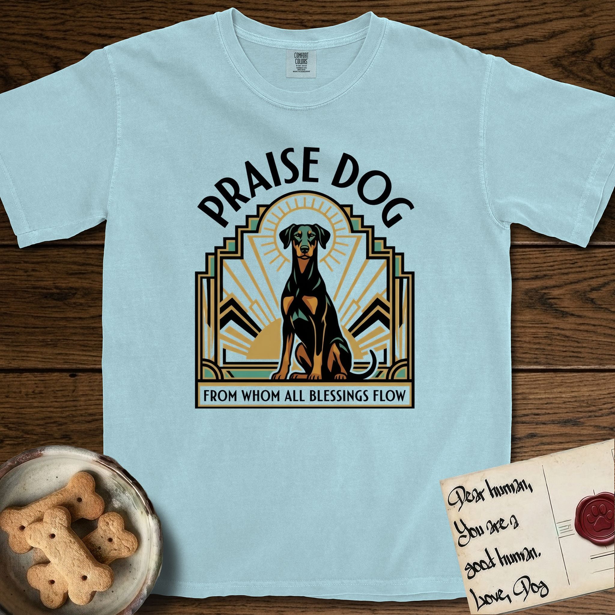 Praise Dog From Whom All Blessings Flow Doberman T-Shirt flat lay in Chambray, featuring an Art Deco emblem of a seated Doberman with natural ears and full tail, framed by geometric gold and jade rays with a sunburst halo and "Praise Dog From Whom All Blessings Flow" lettering.