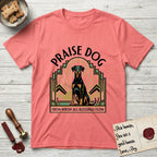 Praise Dog From Whom All Blessings Flow Doberman T-Shirt flat lay in Coral Silk, featuring an Art Deco emblem of a seated Doberman with natural ears and full tail, framed by geometric gold and jade rays with a sunburst halo and "Praise Dog From Whom All Blessings Flow" lettering.