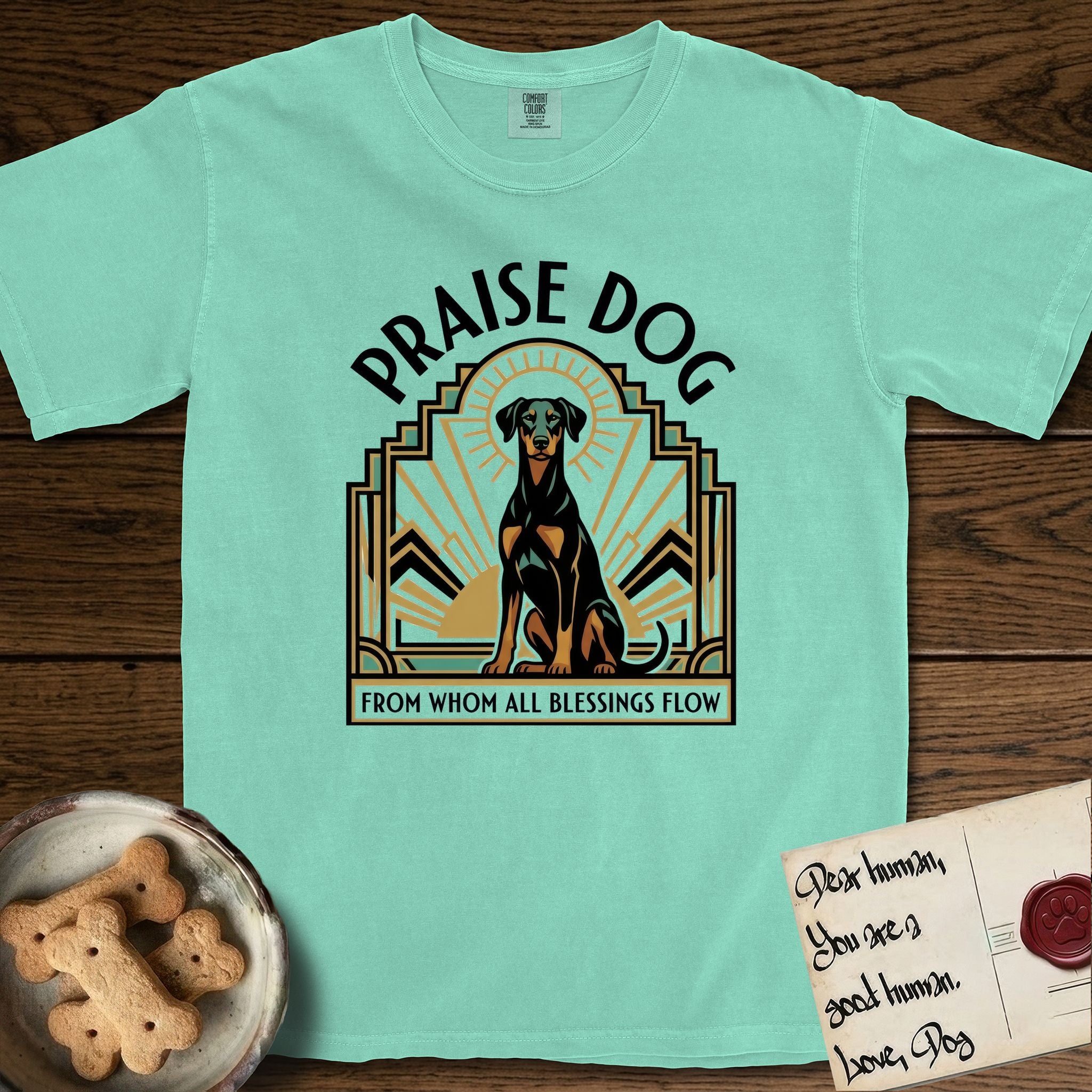 Praise Dog From Whom All Blessings Flow Doberman T-Shirt flat lay in Island Reef, featuring an Art Deco emblem of a seated Doberman with natural ears and full tail, framed by geometric gold and jade rays with a sunburst halo and "Praise Dog From Whom All Blessings Flow" lettering.