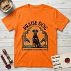 Praise Dog From Whom All Blessings Flow Doberman T-Shirt flat lay in Orange, featuring an Art Deco emblem of a seated Doberman with natural ears and full tail, framed by geometric gold and jade rays with a sunburst halo and "Praise Dog From Whom All Blessings Flow" lettering.