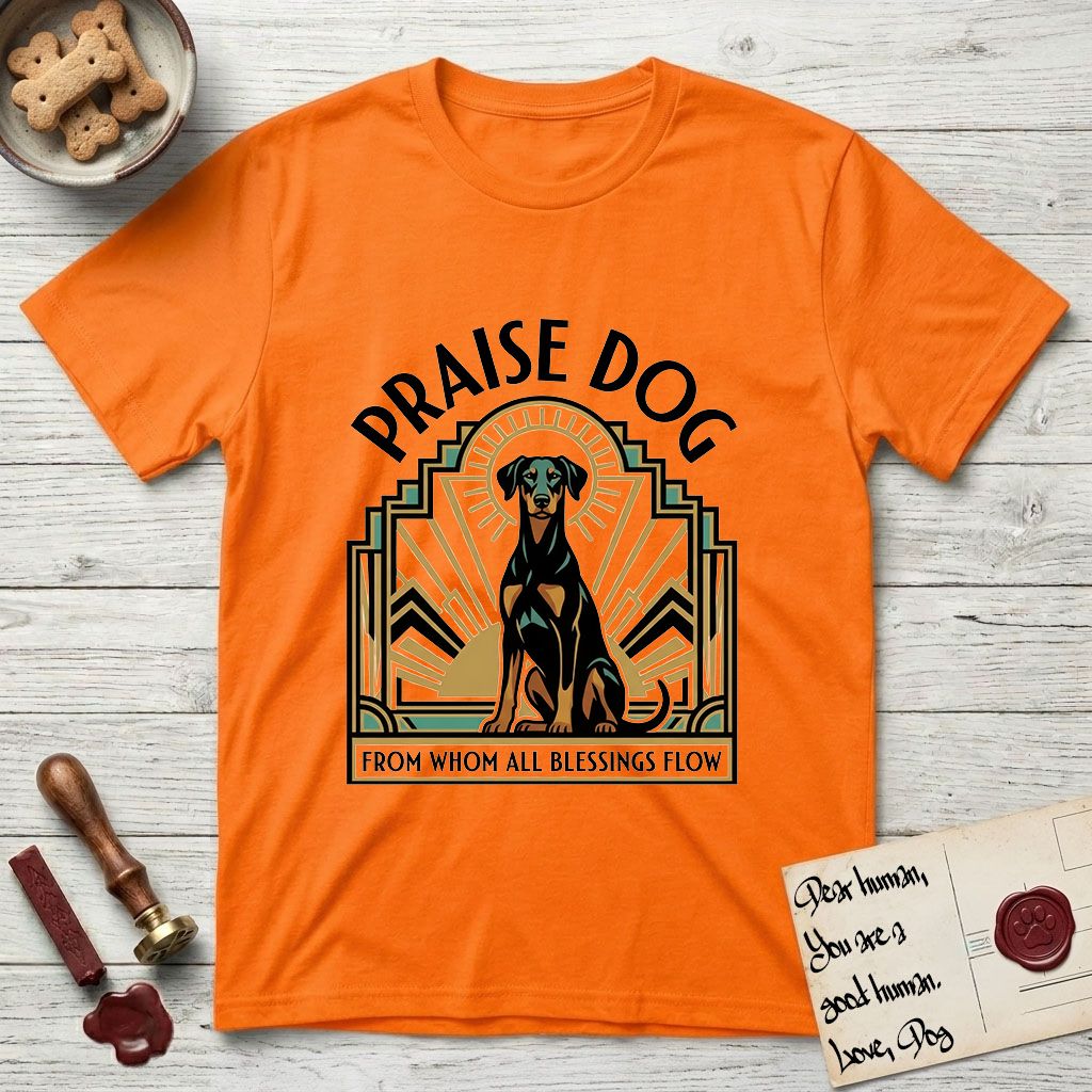 Praise Dog From Whom All Blessings Flow Doberman T-Shirt flat lay in Orange, featuring an Art Deco emblem of a seated Doberman with natural ears and full tail, framed by geometric gold and jade rays with a sunburst halo and "Praise Dog From Whom All Blessings Flow" lettering.