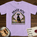 Praise Dog From Whom All Blessings Flow Doberman T-Shirt flat lay in Orchid, featuring an Art Deco emblem of a seated Doberman with natural ears and full tail, framed by geometric gold and jade rays with a sunburst halo and "Praise Dog From Whom All Blessings Flow" lettering.
