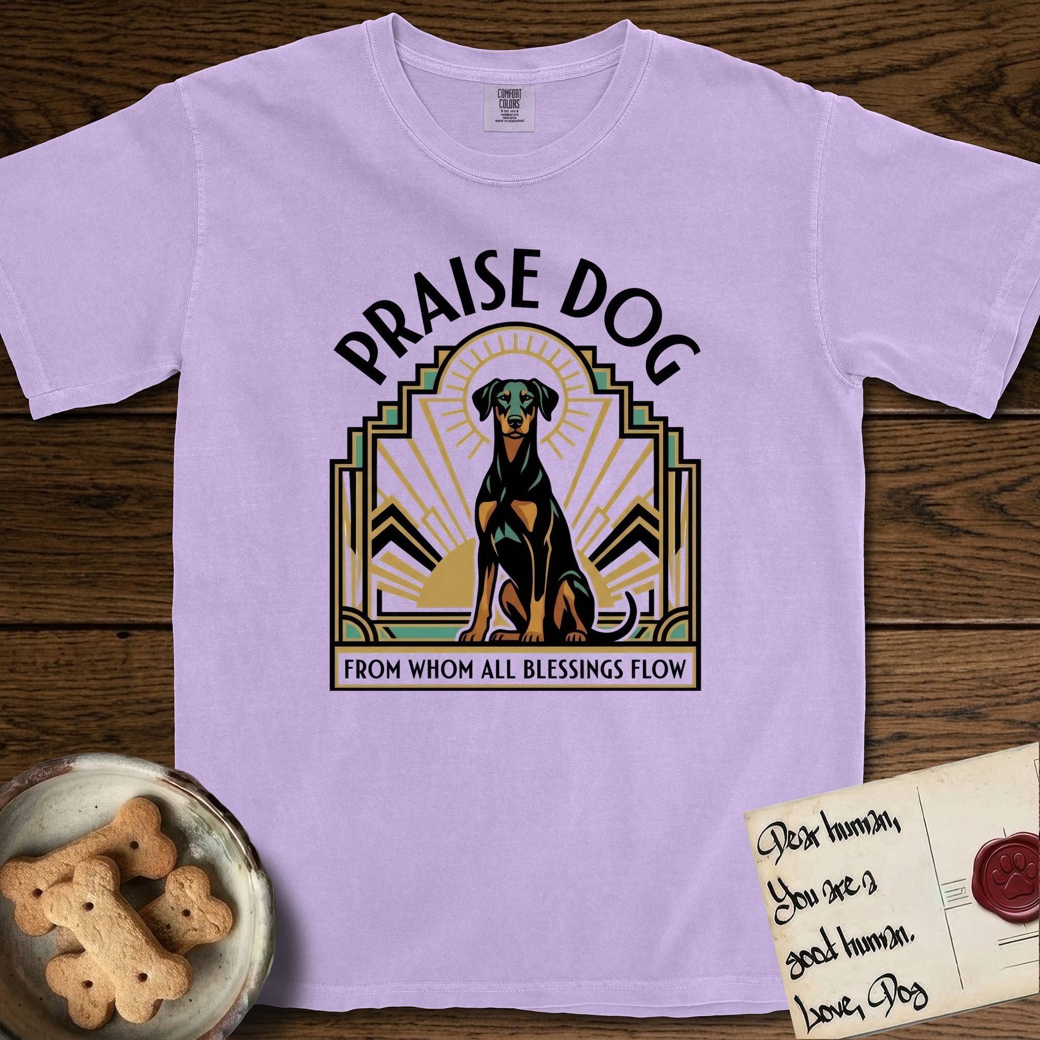 Praise Dog From Whom All Blessings Flow Doberman T-Shirt flat lay in Orchid, featuring an Art Deco emblem of a seated Doberman with natural ears and full tail, framed by geometric gold and jade rays with a sunburst halo and "Praise Dog From Whom All Blessings Flow" lettering.