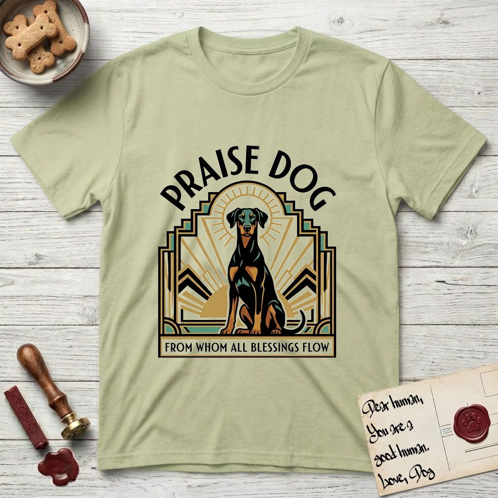 Praise Dog From Whom All Blessings Flow Doberman T-Shirt flat lay in Pistachio, featuring an Art Deco emblem of a seated Doberman with natural ears and full tail, framed by geometric gold and jade rays with a sunburst halo and "Praise Dog From Whom All Blessings Flow" lettering.