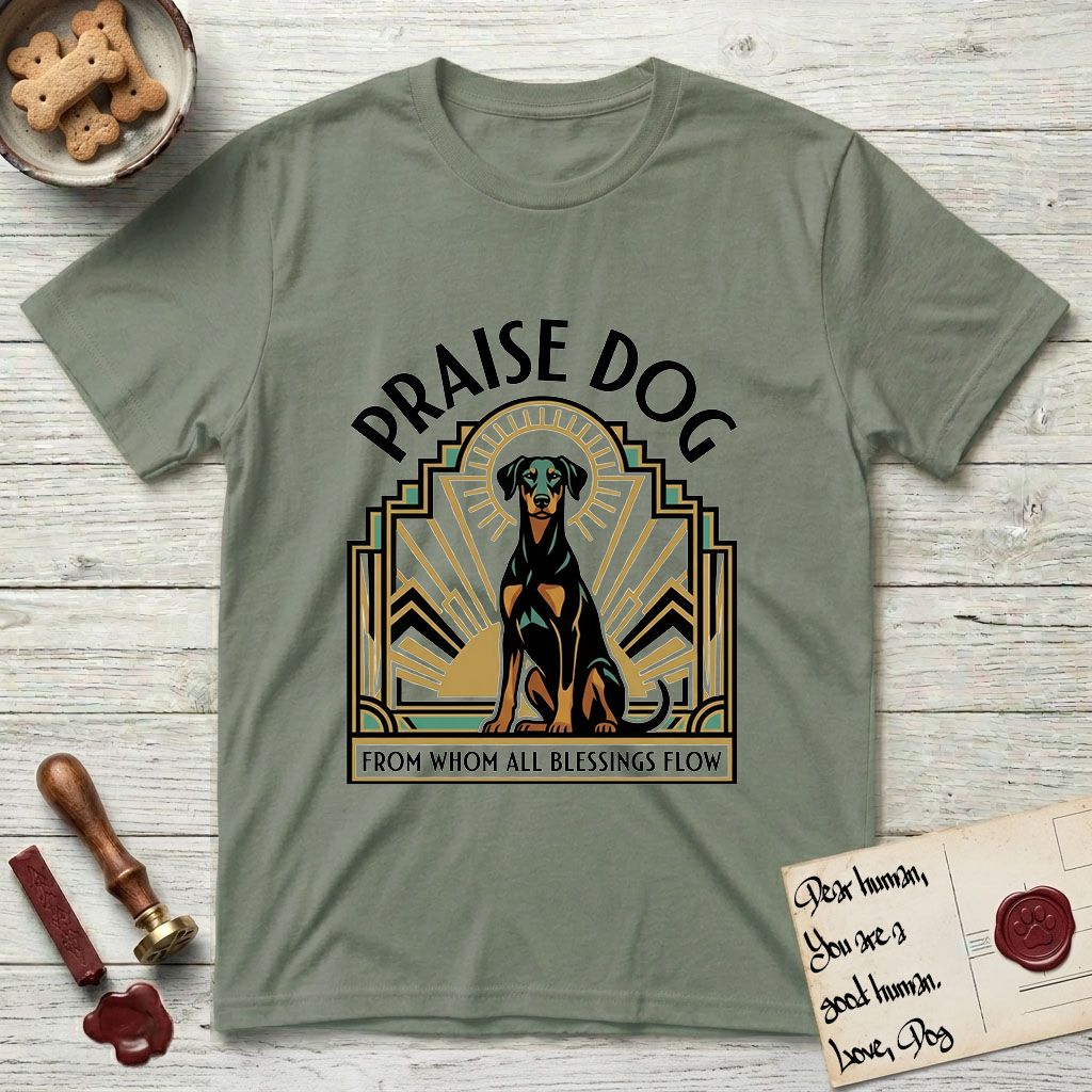 Praise Dog From Whom All Blessings Flow Doberman T-Shirt flat lay in Sage, featuring an Art Deco emblem of a seated Doberman with natural ears and full tail, framed by geometric gold and jade rays with a sunburst halo and "Praise Dog From Whom All Blessings Flow" lettering.
