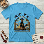 Praise Dog From Whom All Blessings Flow Doberman T-Shirt flat lay in Sky, featuring an Art Deco emblem of a seated Doberman with natural ears and full tail, framed by geometric gold and jade rays with a sunburst halo and "Praise Dog From Whom All Blessings Flow" lettering.