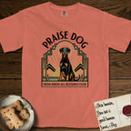Praise Dog From Whom All Blessings Flow Doberman T-Shirt flat lay in Terracotta, featuring an Art Deco emblem of a seated Doberman with natural ears and full tail, framed by geometric gold and jade rays with a sunburst halo and "Praise Dog From Whom All Blessings Flow" lettering.