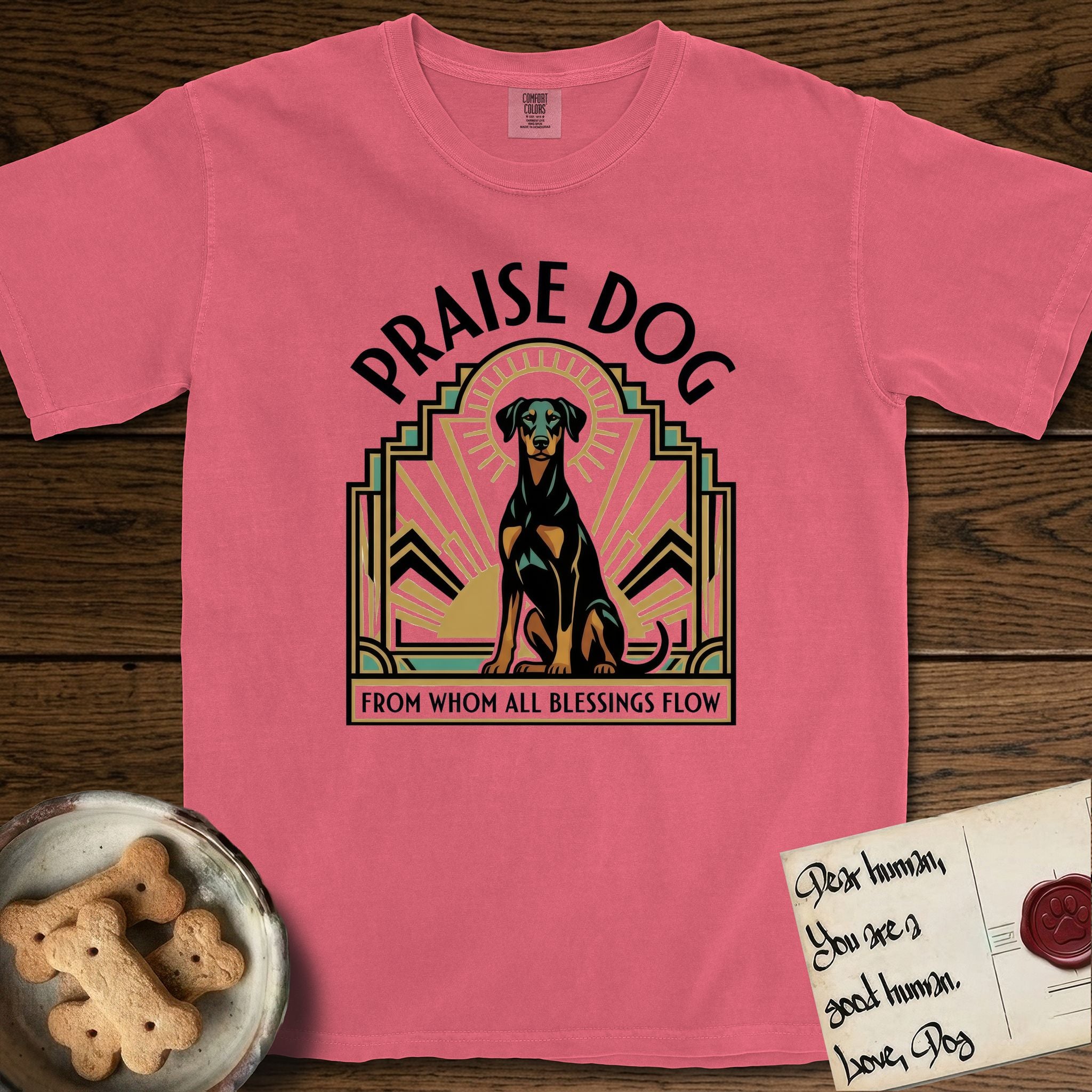 Praise Dog From Whom All Blessings Flow Doberman T-Shirt flat lay in Watermelon, featuring an Art Deco emblem of a seated Doberman with natural ears and full tail, framed by geometric gold and jade rays with a sunburst halo and "Praise Dog From Whom All Blessings Flow" lettering.