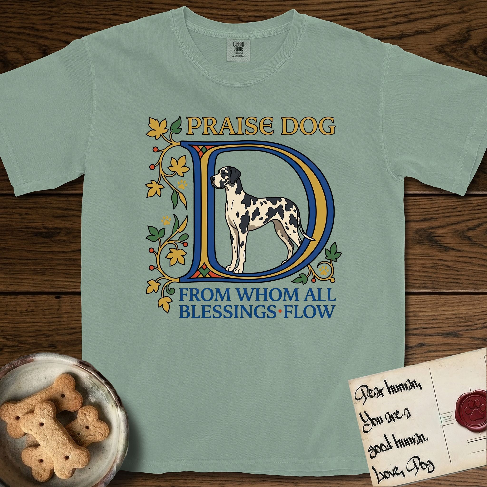 Praise Dog From Whom All Blessings Flow Great Dane T-Shirt flat lay in Bay, featuring an illuminated manuscript capital D in blue and gold with a harlequin Great Dane standing inside the letter and the words Praise Dog From Whom All Blessings Flow.