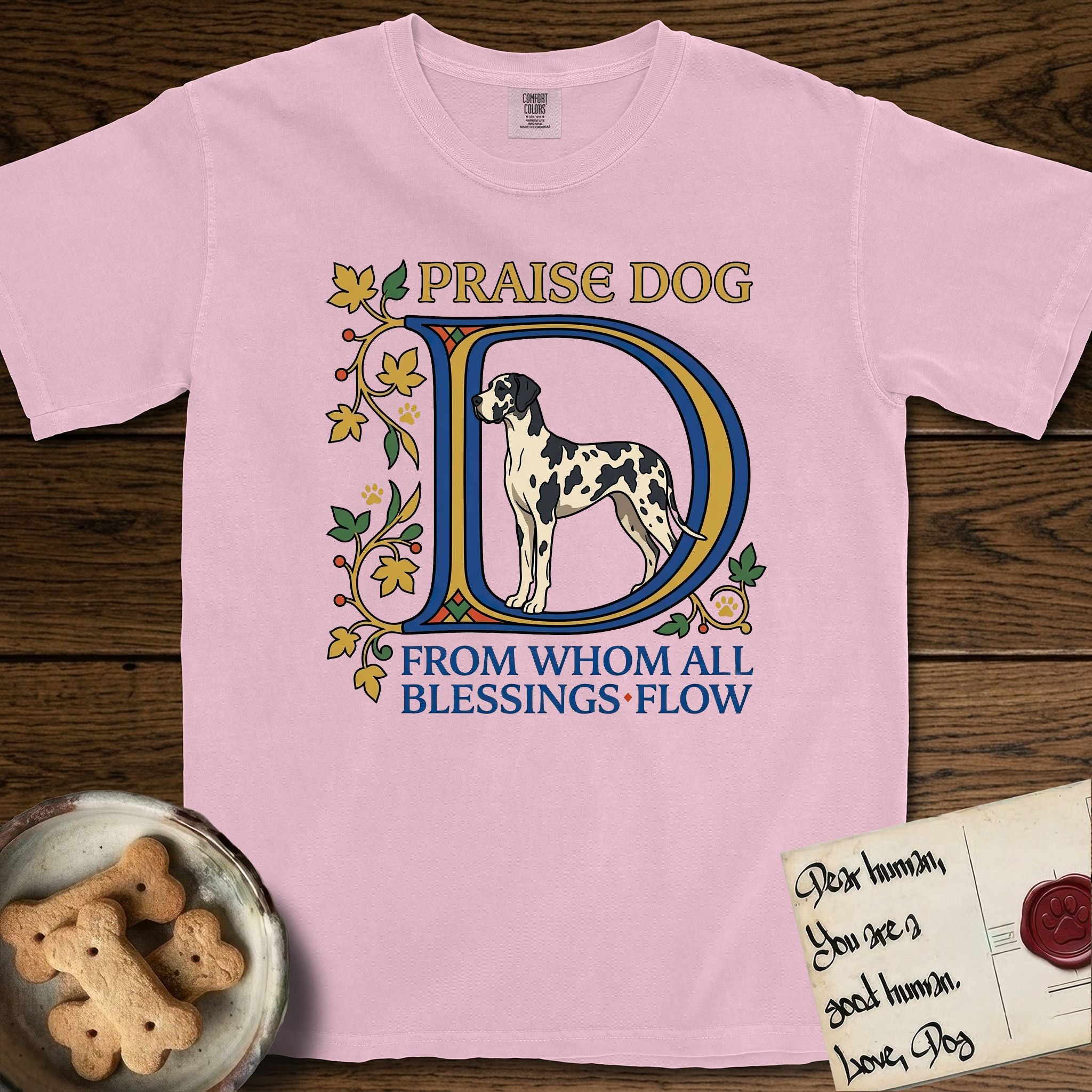 Praise Dog From Whom All Blessings Flow Great Dane T-Shirt flat lay in Blossom, featuring an illuminated manuscript capital D in blue and gold with a harlequin Great Dane standing inside the letter and the words Praise Dog From Whom All Blessings Flow.