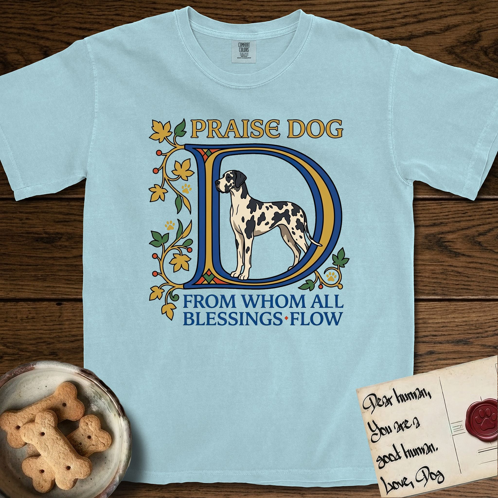 Praise Dog From Whom All Blessings Flow Great Dane T-Shirt flat lay in Chambray, featuring an illuminated manuscript capital D in blue and gold with a harlequin Great Dane standing inside the letter and the words Praise Dog From Whom All Blessings Flow.