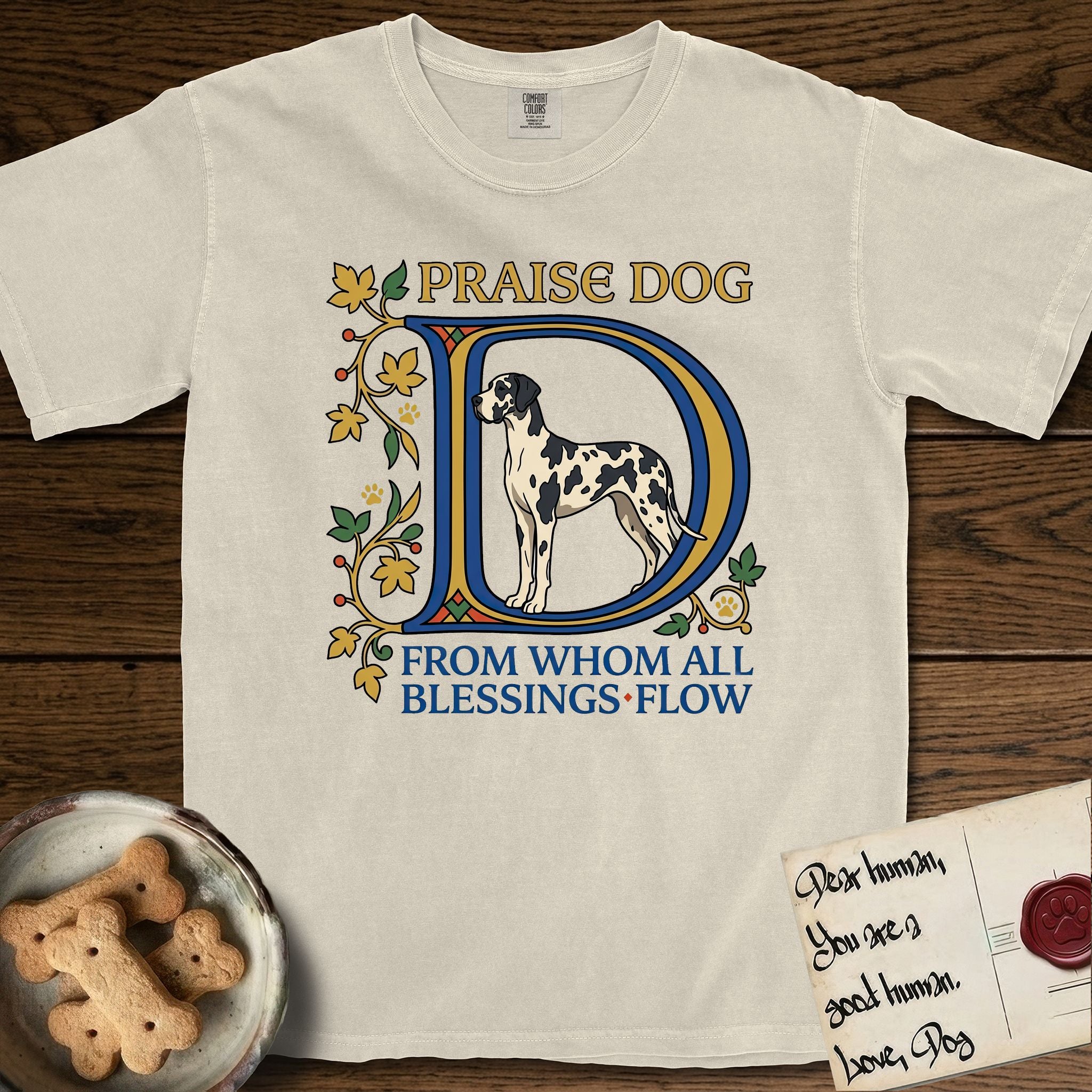 Praise Dog From Whom All Blessings Flow Great Dane T-Shirt flat lay in Ivory, featuring an illuminated manuscript capital D in blue and gold with a harlequin Great Dane standing inside the letter and the words Praise Dog From Whom All Blessings Flow.