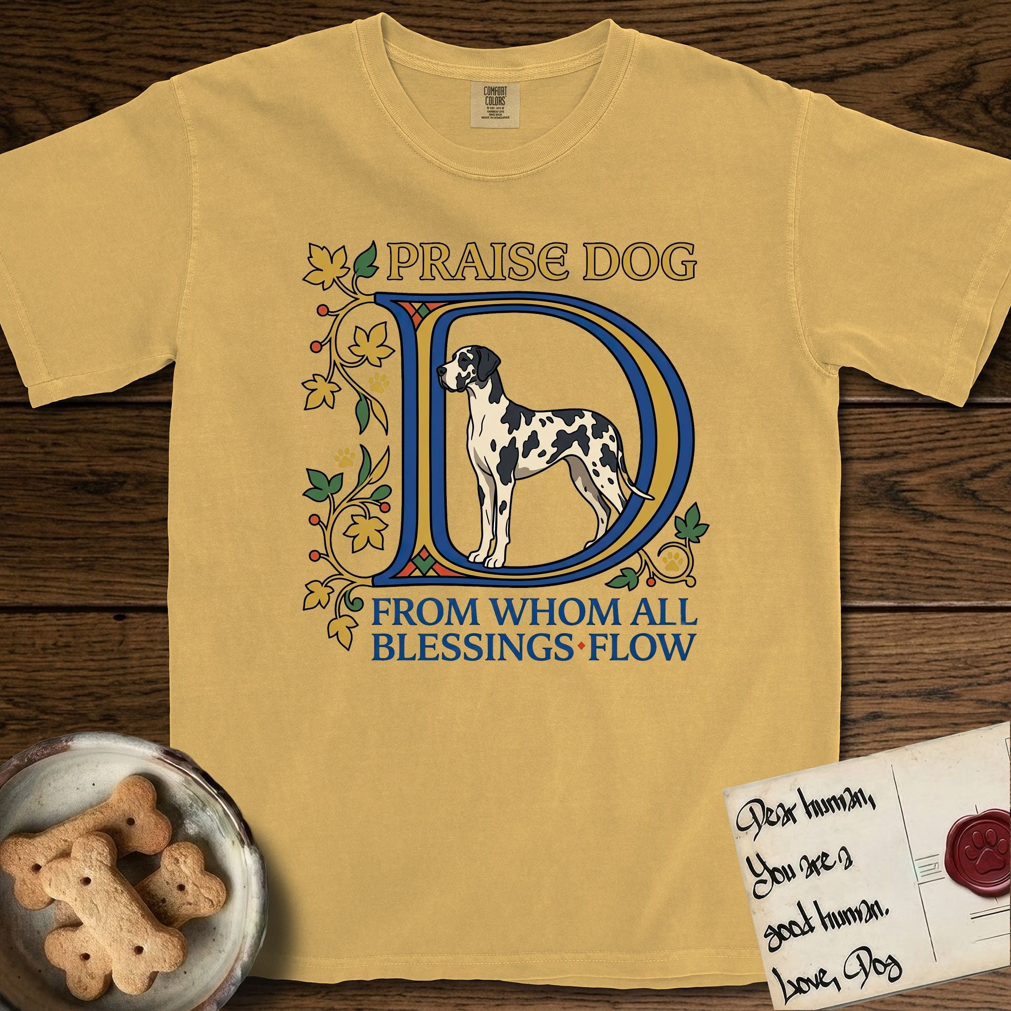 Praise Dog From Whom All Blessings Flow Great Dane T-Shirt flat lay in Mustard, featuring an illuminated manuscript capital D in blue and gold with a harlequin Great Dane standing inside the letter and the words Praise Dog From Whom All Blessings Flow.