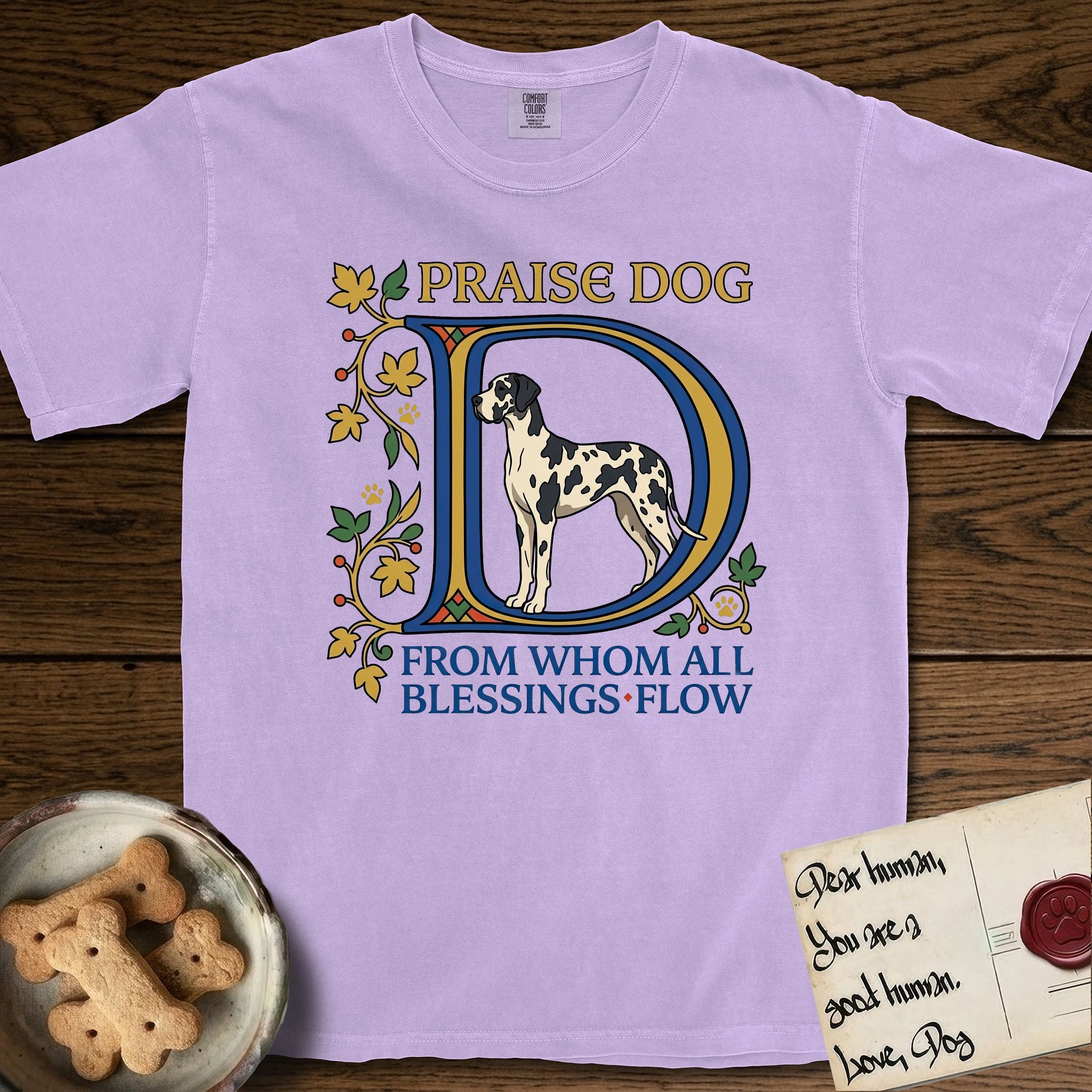 Praise Dog From Whom All Blessings Flow Great Dane T-Shirt flat lay in Orchid, featuring an illuminated manuscript capital D in blue and gold with a harlequin Great Dane standing inside the letter and the words Praise Dog From Whom All Blessings Flow.