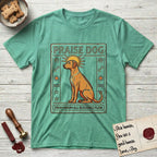 Praise Dog From Whom All Blessings Flow Rhodesian Ridgeback T-Shirt flat lay in Heather Irish Green, featuring an ancient Egyptian carved-relief emblem of a Rhodesian Ridgeback in side profile with a visible spine ridge, a rayed golden sun halo, hieroglyph glyph columns, and a ragged stone border with "Praise Dog From Whom All Blessings Flow" lettering.