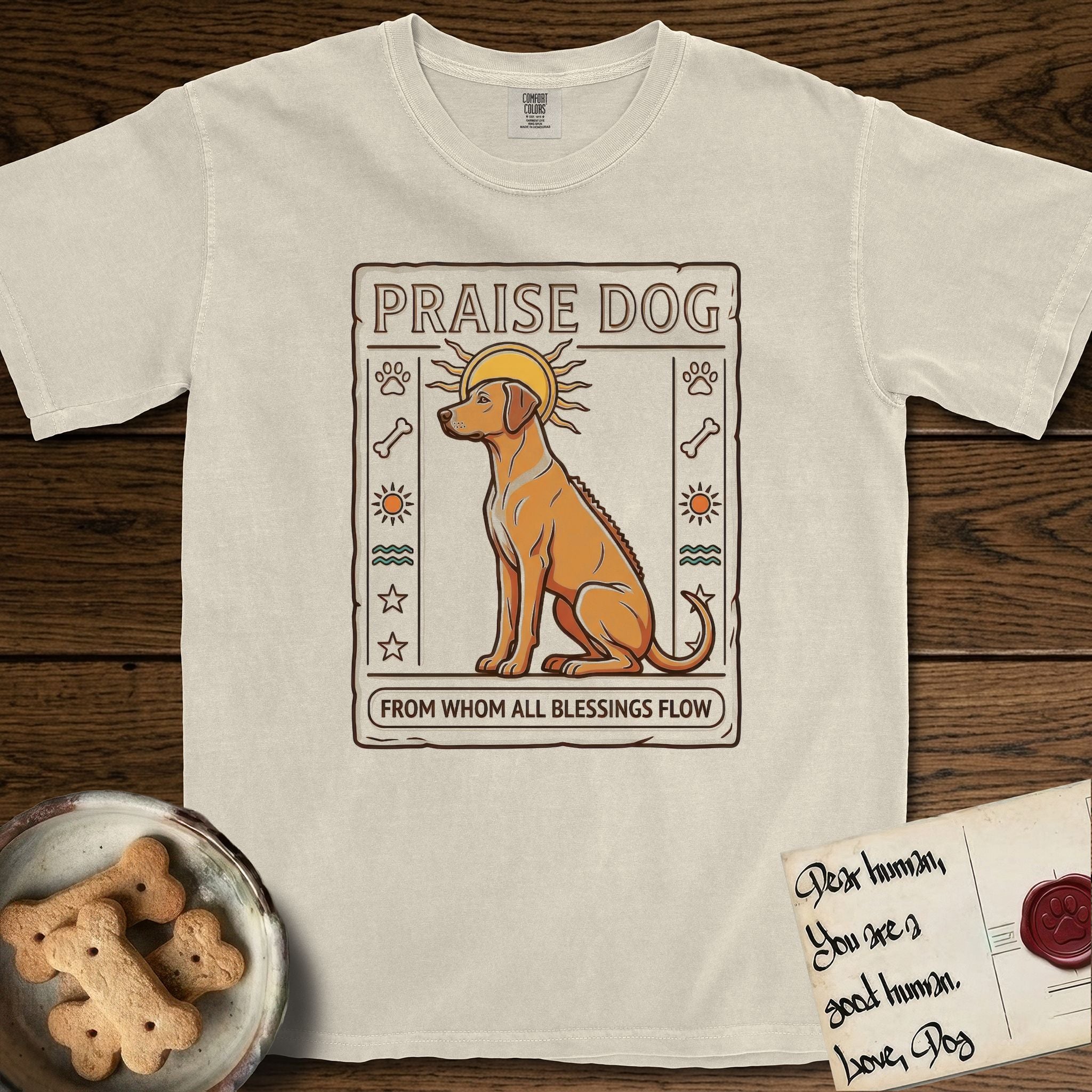 Praise Dog From Whom All Blessings Flow Rhodesian Ridgeback T-Shirt flat lay in Ivory, featuring an ancient Egyptian carved-relief emblem of a Rhodesian Ridgeback in side profile with a visible spine ridge, a rayed golden sun halo, hieroglyph glyph columns, and a ragged stone border with "Praise Dog From Whom All Blessings Flow" lettering.