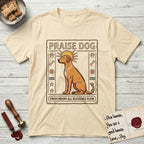 Praise Dog From Whom All Blessings Flow Rhodesian Ridgeback T-Shirt flat lay in Natural, featuring an ancient Egyptian carved-relief emblem of a Rhodesian Ridgeback in side profile with a visible spine ridge, a rayed golden sun halo, hieroglyph glyph columns, and a ragged stone border with "Praise Dog From Whom All Blessings Flow" lettering.