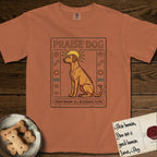 Praise Dog From Whom All Blessings Flow Rhodesian Ridgeback T-Shirt flat lay in Yam, featuring an ancient Egyptian carved-relief emblem of a Rhodesian Ridgeback in side profile with a visible spine ridge, a rayed golden sun halo, hieroglyph glyph columns, and a ragged stone border with "Praise Dog From Whom All Blessings Flow" lettering.