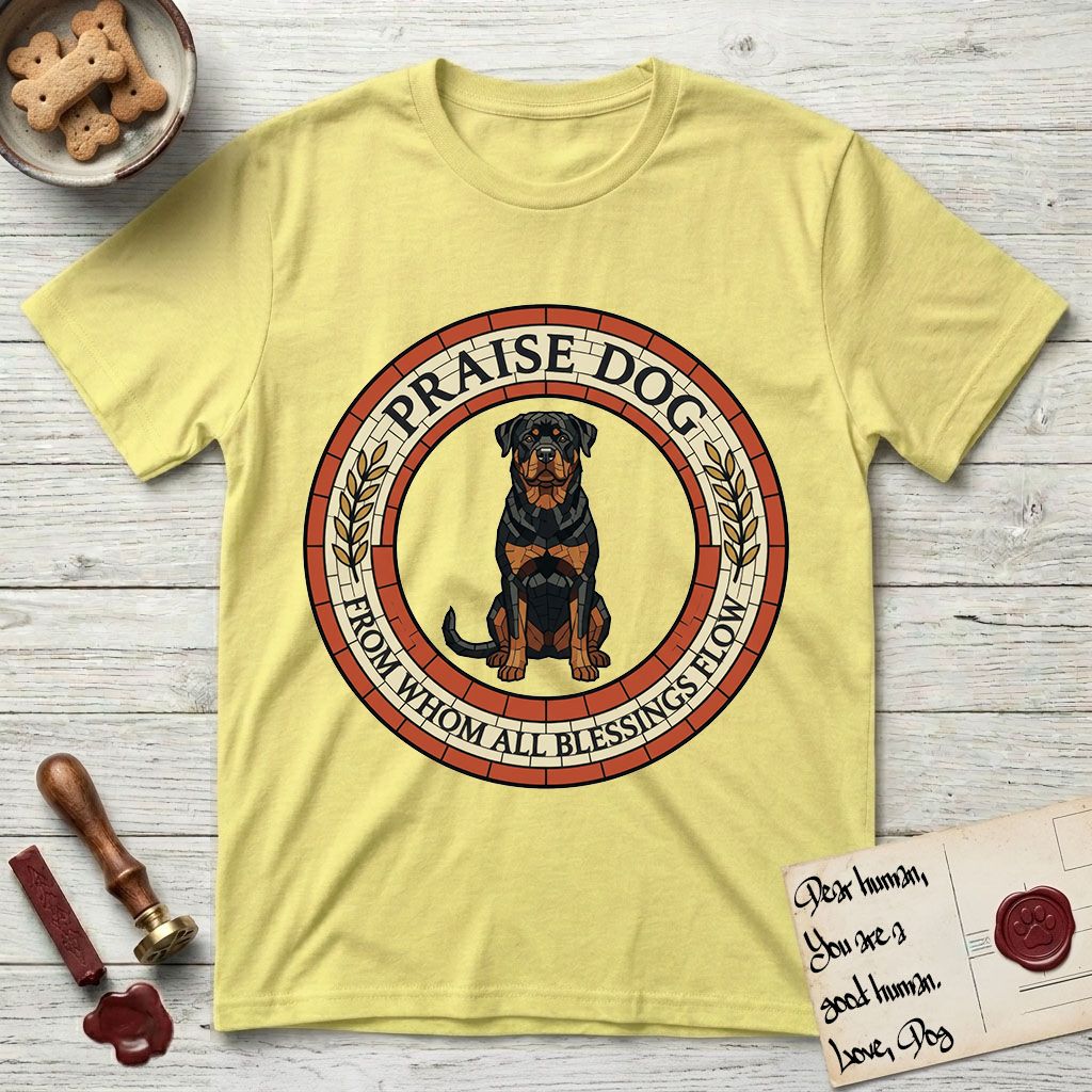 Praise Dog From Whom All Blessings Flow Rottweiler T-Shirt flat lay in Cornsilk, featuring a Roman mosaic ring medallion of a seated Rottweiler with natural ears and a full undocked tail, built from terracotta and mahogany tiles and flanked by laurel branches, with "Praise Dog From Whom All Blessings Flow" lettering.