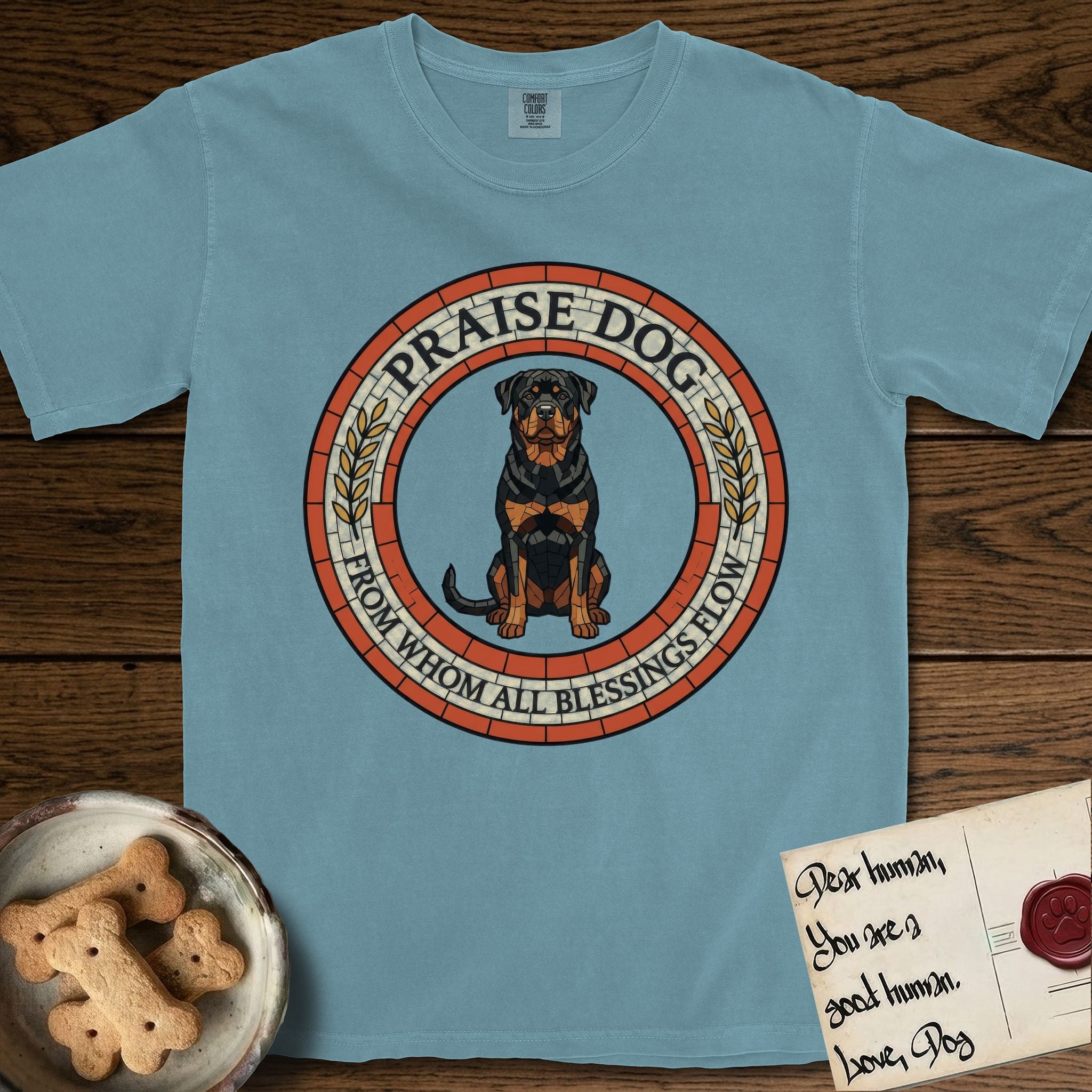 Praise Dog From Whom All Blessings Flow Rottweiler T-Shirt flat lay in Ice Blue, featuring a Roman mosaic ring medallion of a seated Rottweiler with natural ears and a full undocked tail, built from terracotta and mahogany tiles and flanked by laurel branches, with "Praise Dog From Whom All Blessings Flow" lettering.
