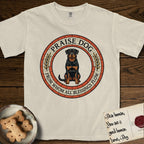 Praise Dog From Whom All Blessings Flow Rottweiler T-Shirt flat lay in Ivory, featuring a Roman mosaic ring medallion of a seated Rottweiler with natural ears and a full undocked tail, built from terracotta and mahogany tiles and flanked by laurel branches, with "Praise Dog From Whom All Blessings Flow" lettering.