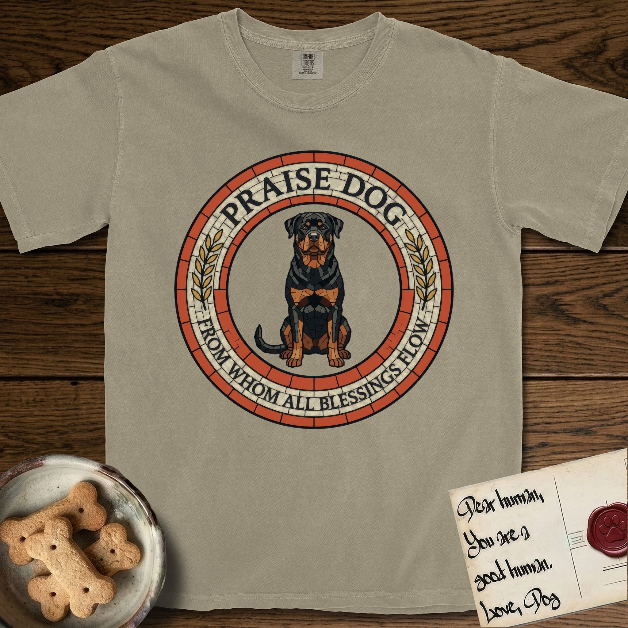 Praise Dog From Whom All Blessings Flow Rottweiler T-Shirt flat lay in Khaki, featuring a Roman mosaic ring medallion of a seated Rottweiler with natural ears and a full undocked tail, built from terracotta and mahogany tiles and flanked by laurel branches, with "Praise Dog From Whom All Blessings Flow" lettering.