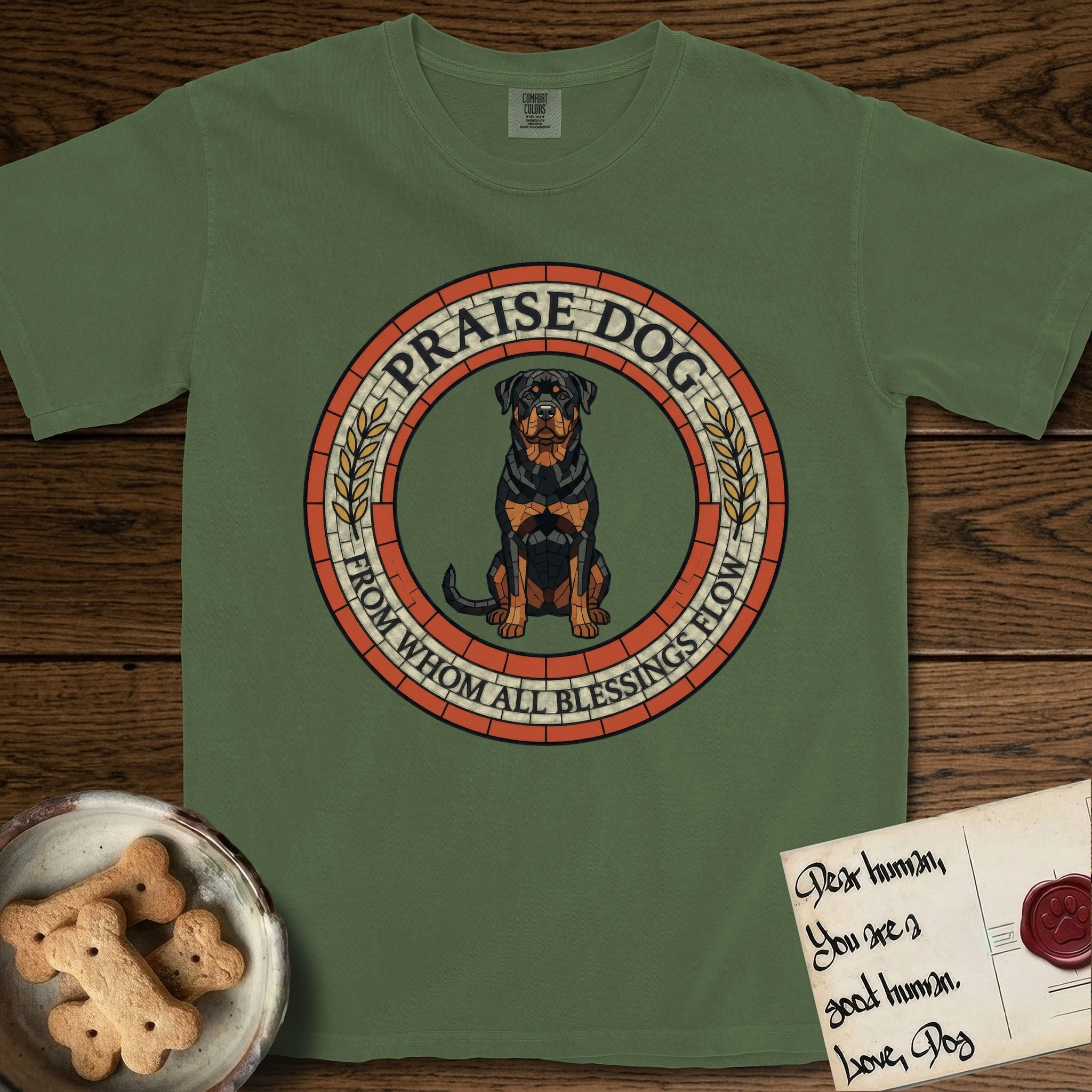 Praise Dog From Whom All Blessings Flow Rottweiler T-Shirt flat lay in Moss, featuring a Roman mosaic ring medallion of a seated Rottweiler with natural ears and a full undocked tail, built from terracotta and mahogany tiles and flanked by laurel branches, with "Praise Dog From Whom All Blessings Flow" lettering.