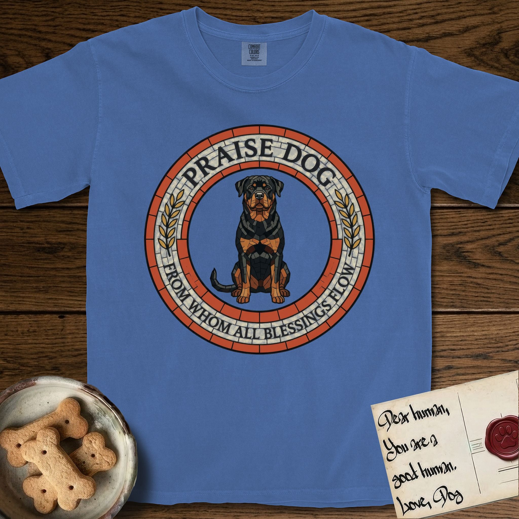 Praise Dog From Whom All Blessings Flow Rottweiler T-Shirt flat lay in Mystic Blue, featuring a Roman mosaic ring medallion of a seated Rottweiler with natural ears and a full undocked tail, built from terracotta and mahogany tiles and flanked by laurel branches, with "Praise Dog From Whom All Blessings Flow" lettering.