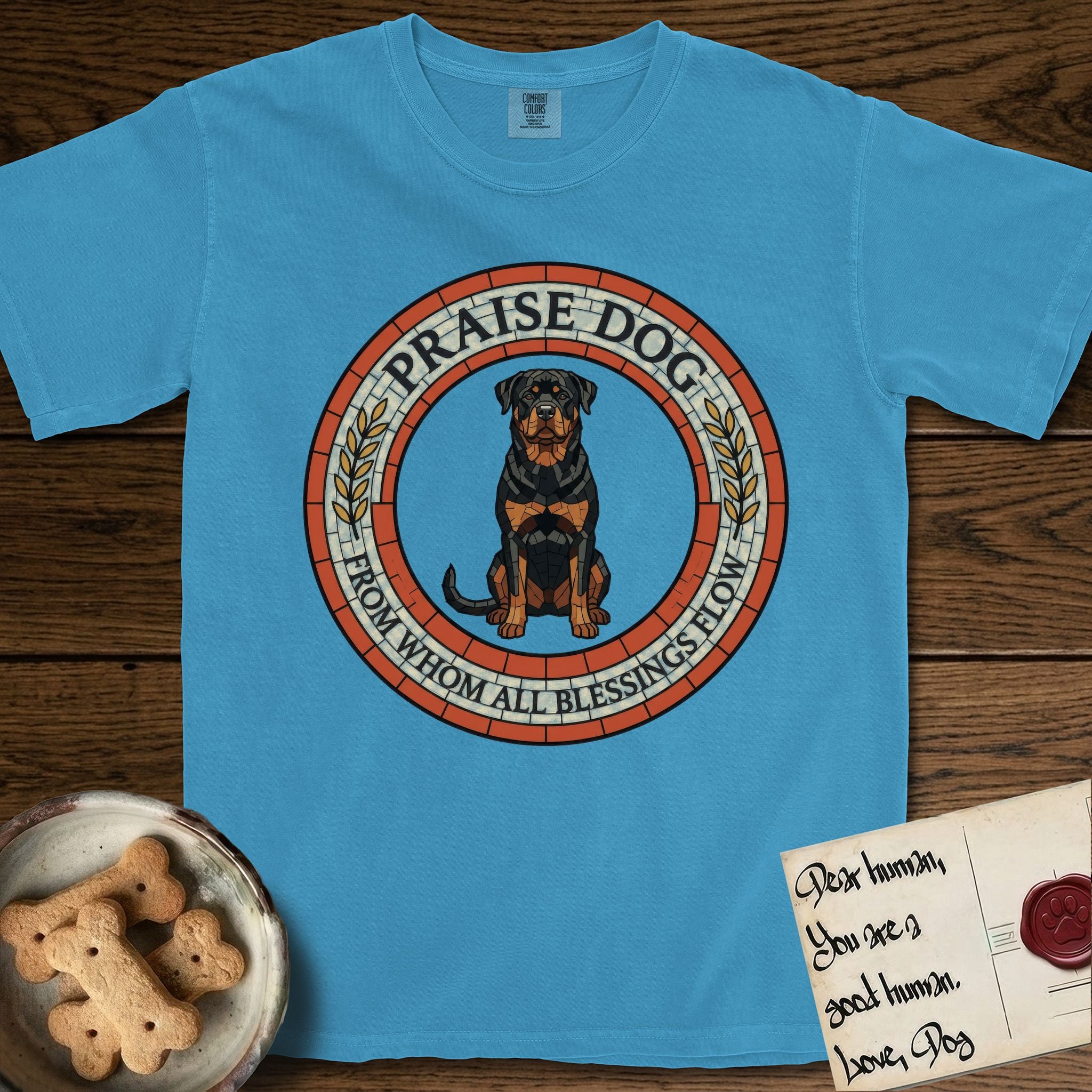 Praise Dog From Whom All Blessings Flow Rottweiler T-Shirt flat lay in Royal Caribe, featuring a Roman mosaic ring medallion of a seated Rottweiler with natural ears and a full undocked tail, built from terracotta and mahogany tiles and flanked by laurel branches, with "Praise Dog From Whom All Blessings Flow" lettering.
