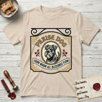 Praise Dog From Whom All Blessings Flow Schnauzer T-Shirt flat lay in Sand, featuring a vintage tavern sign with a bearded Schnauzer portrait in a gilded oval, hops sprigs, and a ribbon banner, with the words Praise Dog From Whom All Blessings Flow.