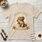 Praise Dog From Whom All Blessings Flow Shih Tzu T-Shirt flat lay in Sand, featuring an old-master devotional oil painting of a Shih Tzu seated with one paw raised in blessing, a floating gold halo and streaming light, a shower of small gold blessings, and gilt "Praise Dog From Whom All Blessings Flow" lettering on a ribbon scroll.