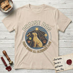 Praise Dog From Whom All Blessings Flow Yellow Lab T-Shirt flat lay in Sand, featuring a warm vintage circular emblem with an upward-gazing Yellow Lab, a sunburst, and a devotional border of small hearts and stars.