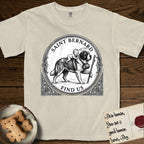 Patron Saint Medallion Saint Bernard T-Shirt flat lay in Ivory, featuring a vintage engraving coin medallion of a haloed Saint Bernard carrying a child across a mountain pass, with the petition FIND US.